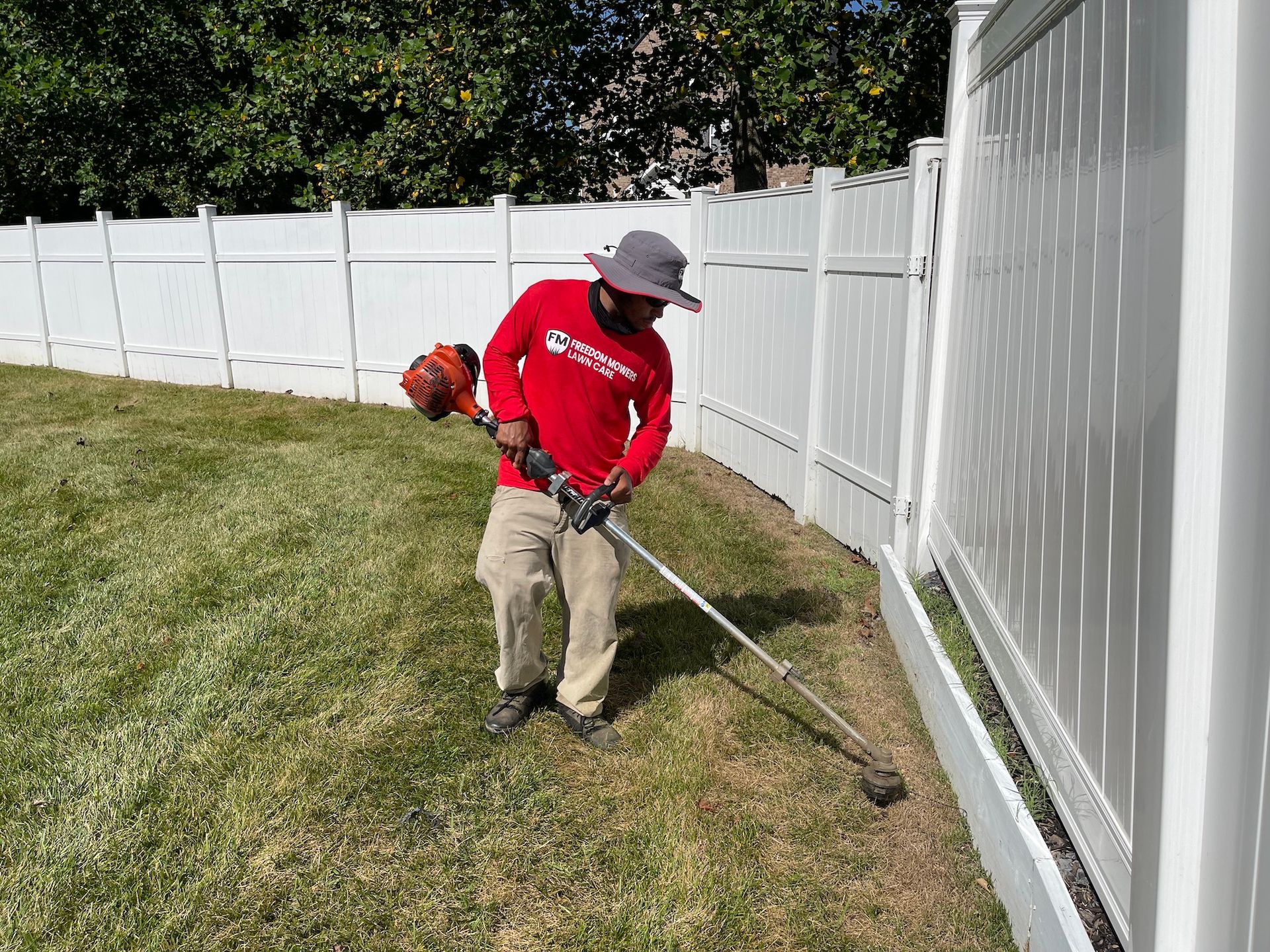 A man is mowing the grass in front of a white fence.