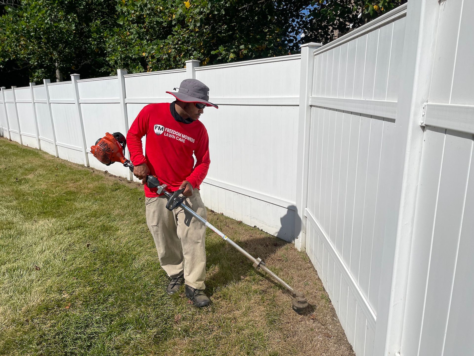 A man is using a lawn mower to cut the grass next to a white fence.
