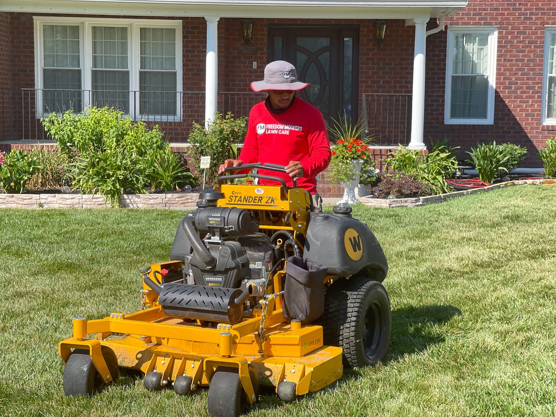 A man is riding a yellow lawn mower on a lush green lawn in front of a brick house.