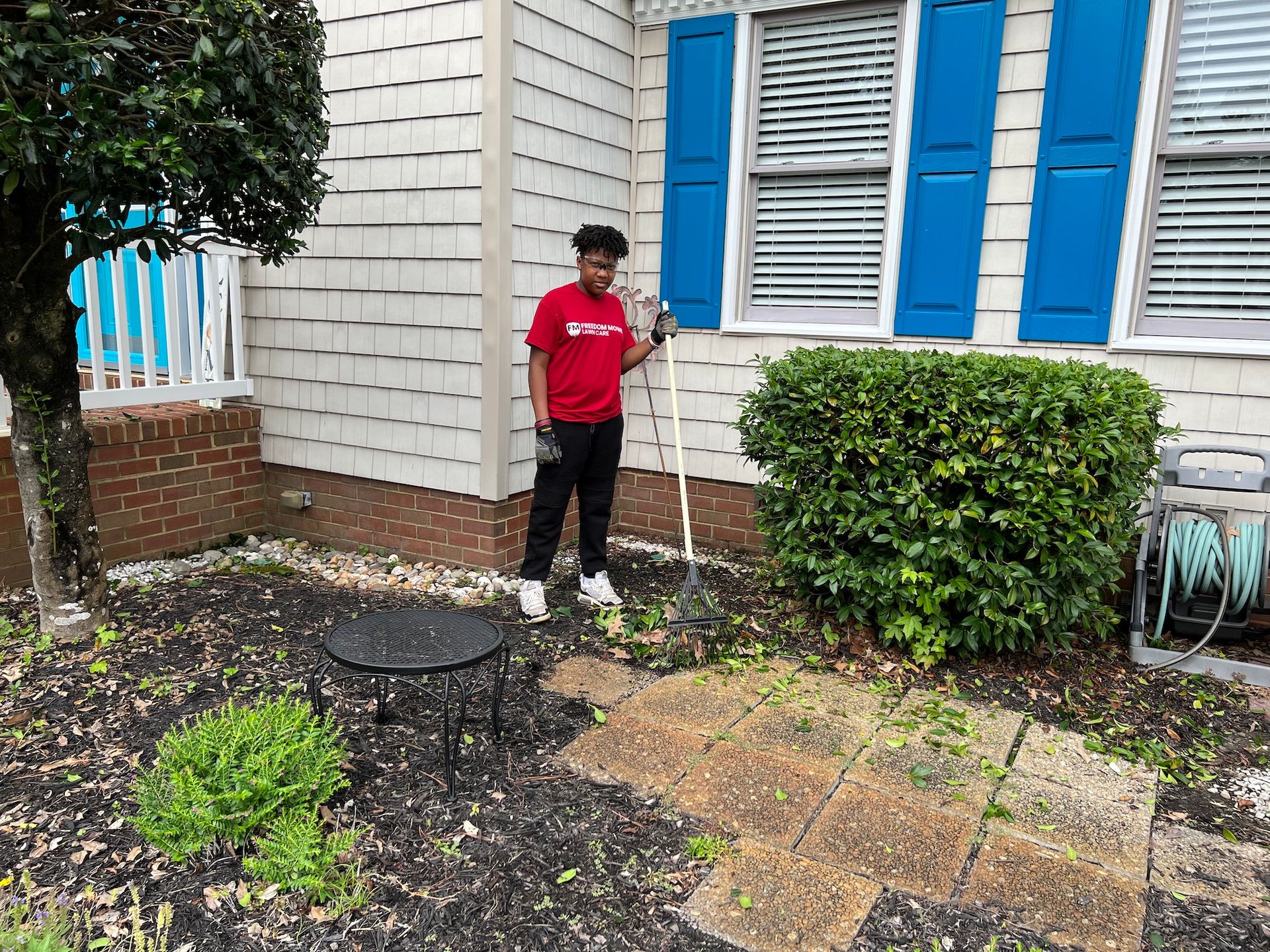 A man in a red shirt is standing in front of a house with blue shutters.