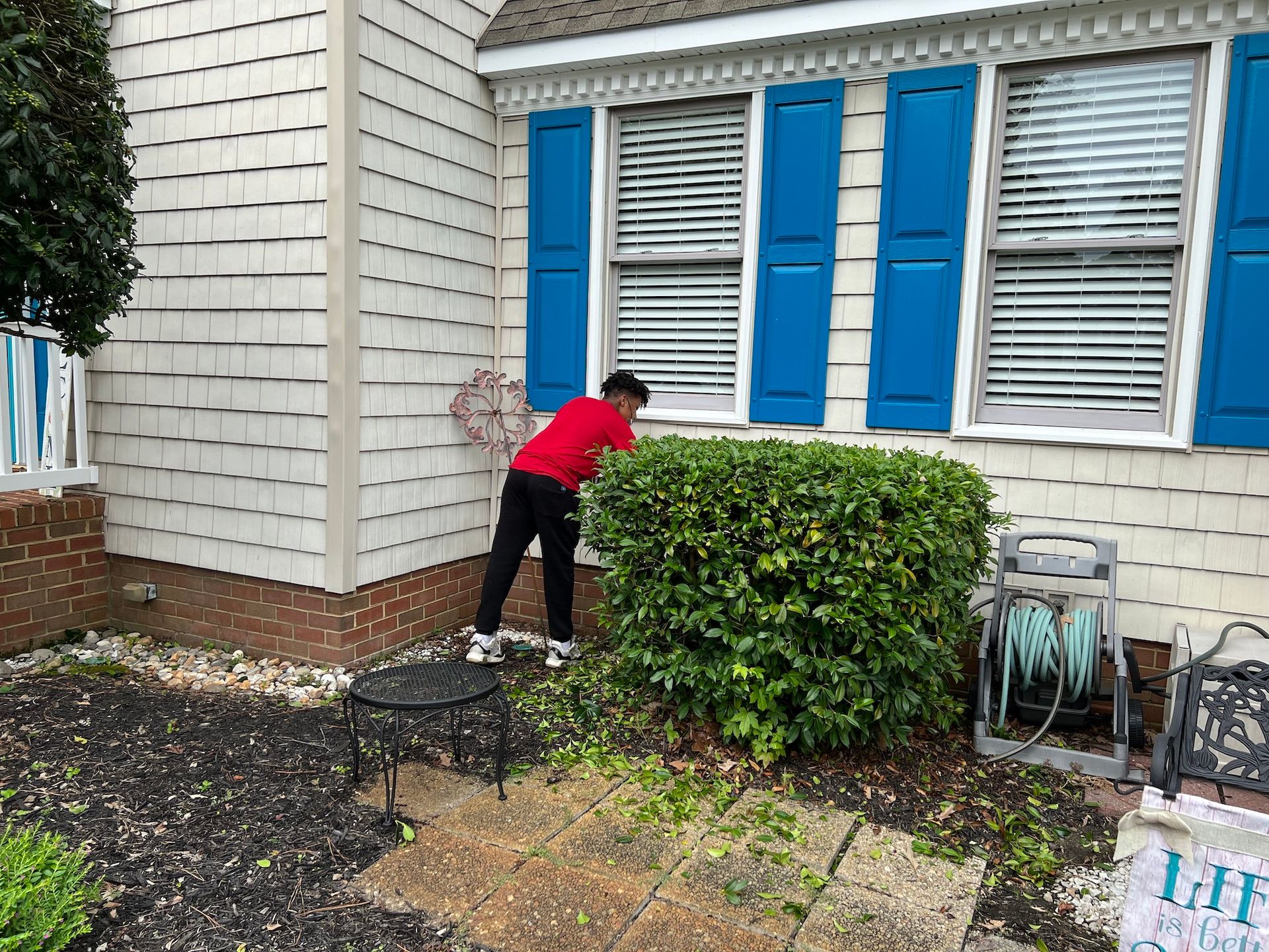 A man in a red shirt is cutting a bush in front of a house with blue shutters.