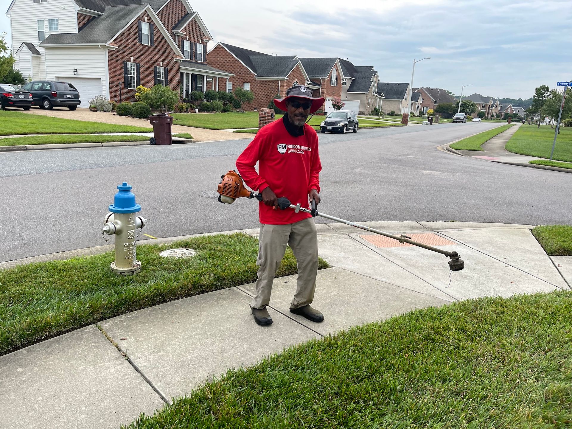 A man is standing on a sidewalk cutting grass with a lawn mower.