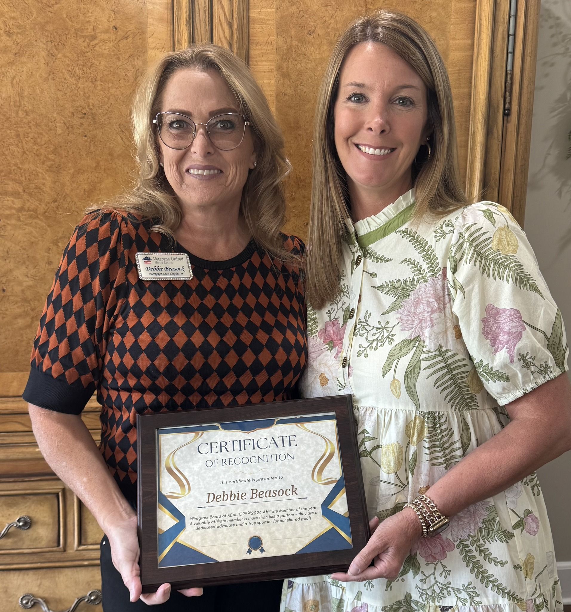 Two women holding a framed certificate in front of a wooden cabinet. One wears a patterned top, the other a floral dress.