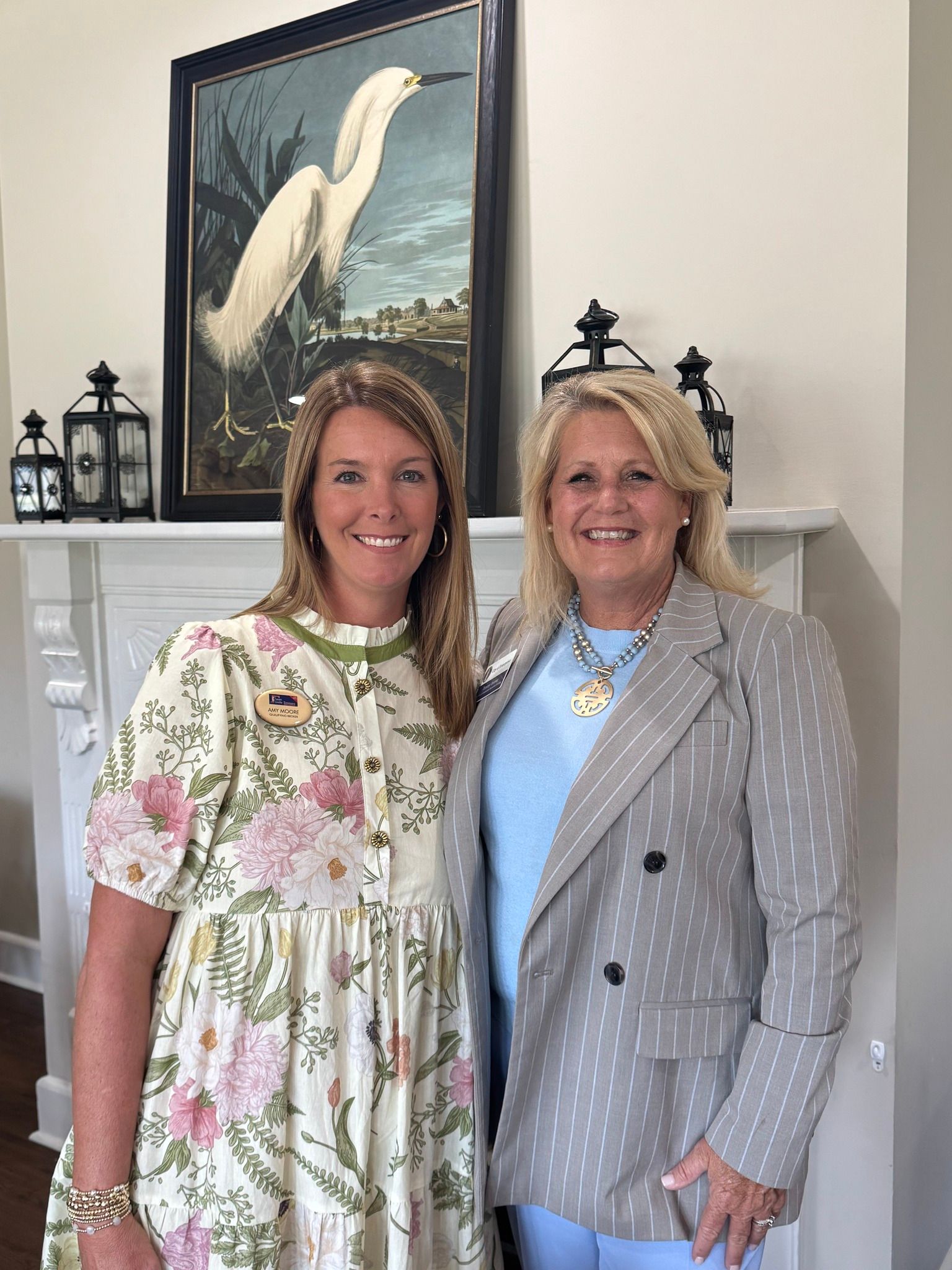 Two women smiling by a fireplace, one in a floral dress, the other in a blazer.