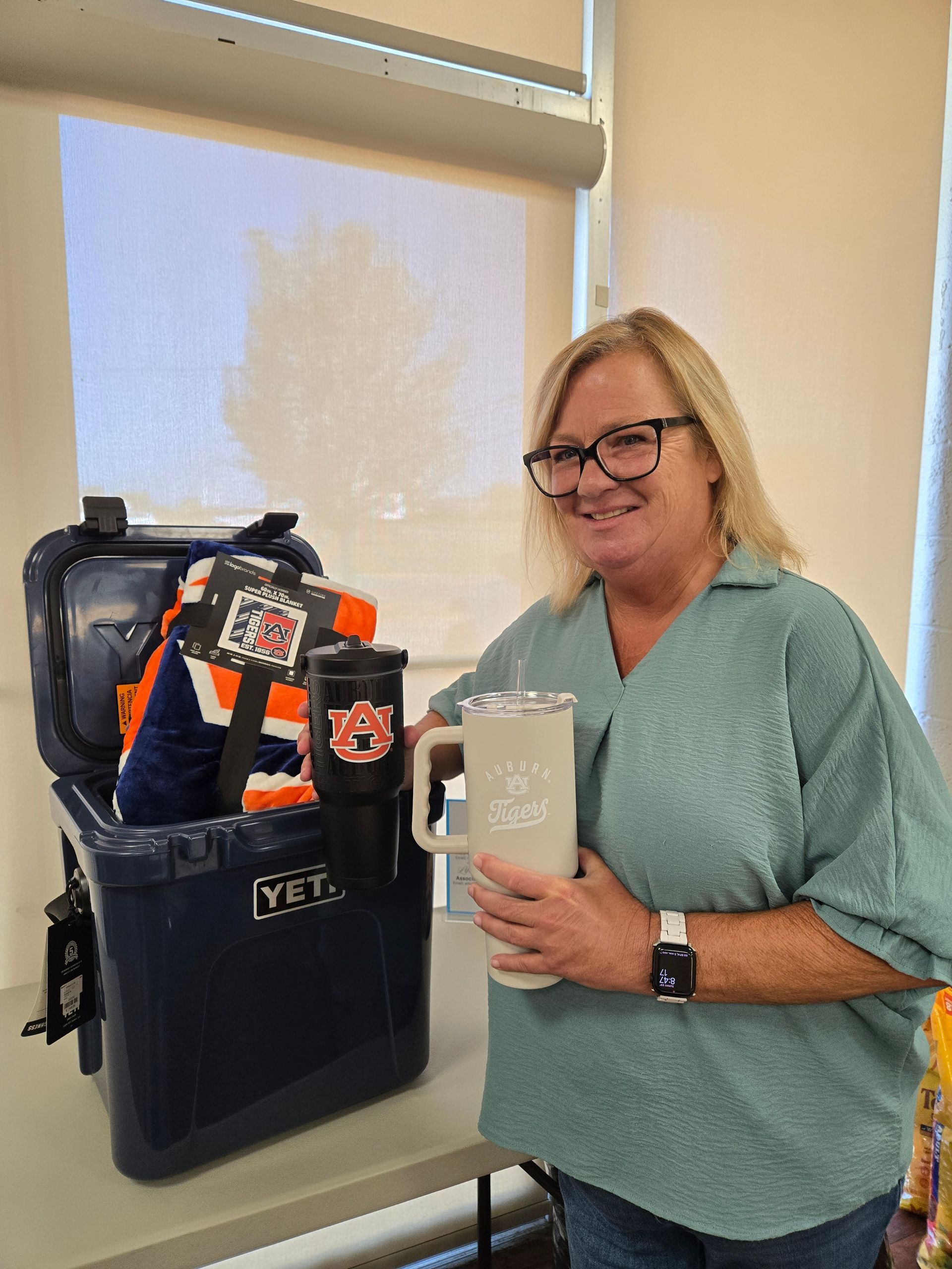 Woman smiles, holding a large mug. Next to her is a navy Yeti cooler filled with Auburn University merchandise.