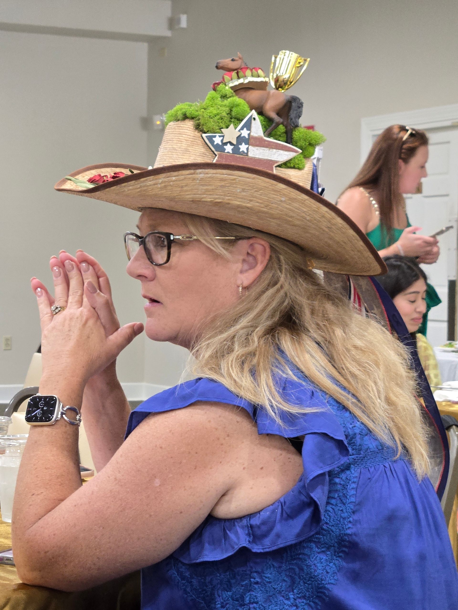 Woman with decorated cowboy hat, hands clasped, wearing glasses and a blue dress, sitting indoors.