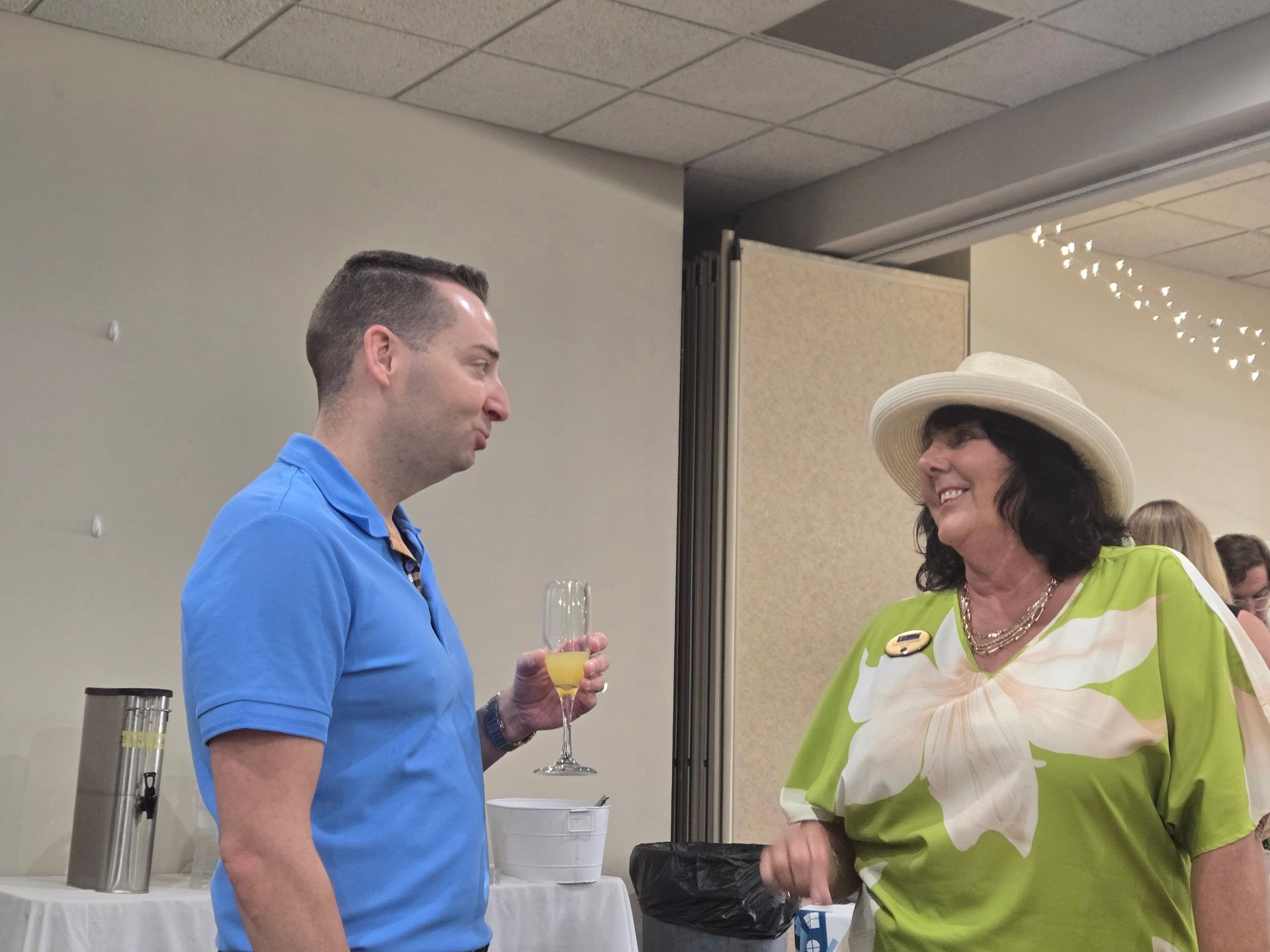 Man in blue shirt and woman in green floral shirt talking, holding drinks at an event.