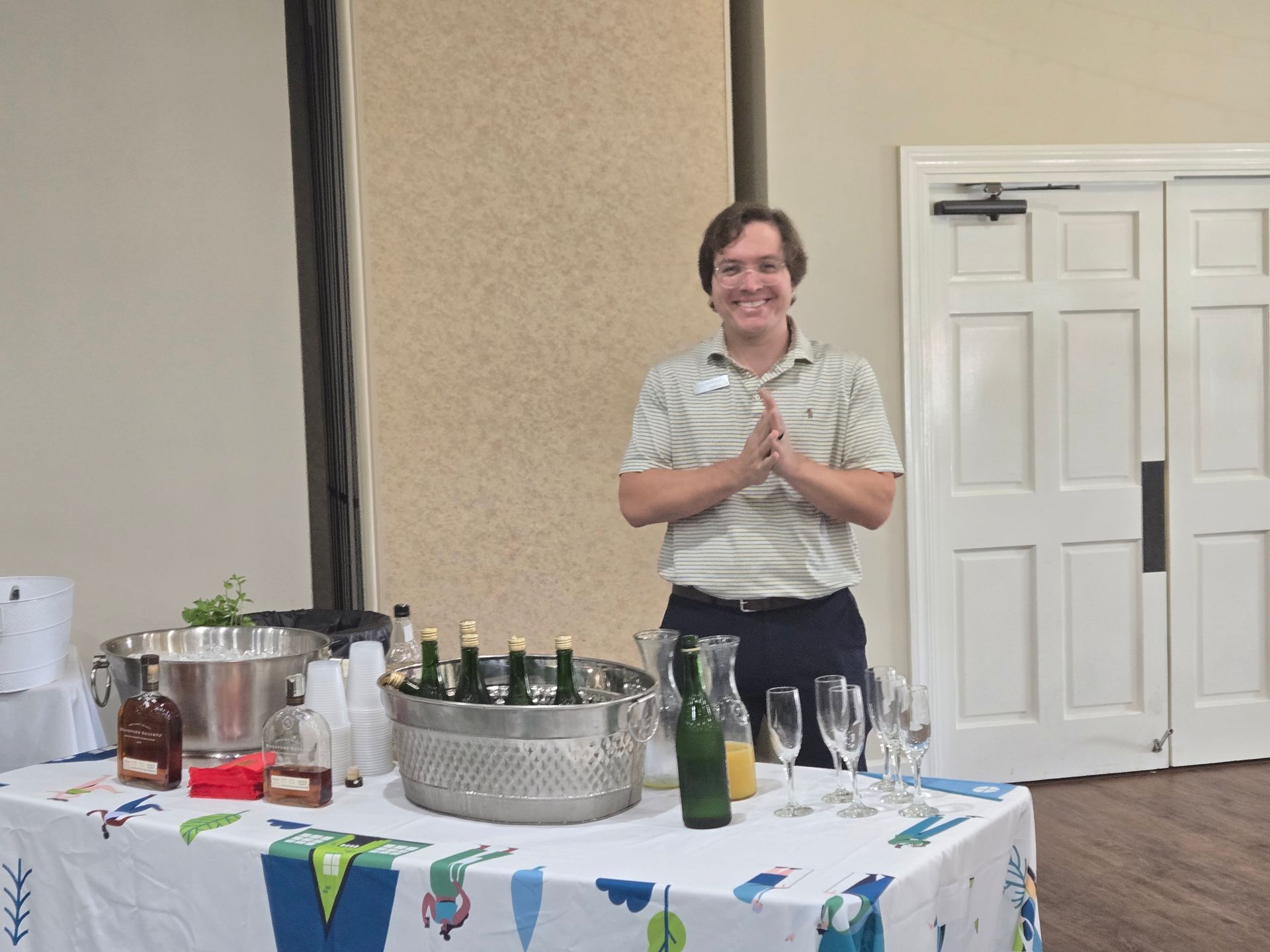 Person standing behind a table with drinks, smiling with hands clasped. Champagne and juice glasses on the table.