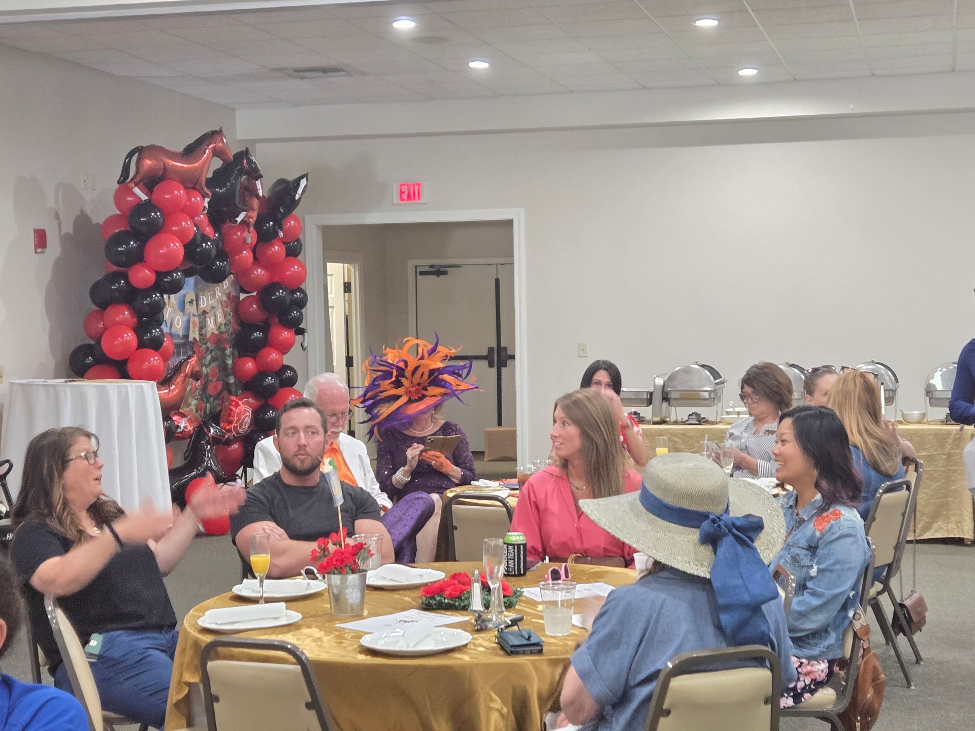 Guests in a room with hats, food, and balloons, sitting at tables, conversing and enjoying the event.