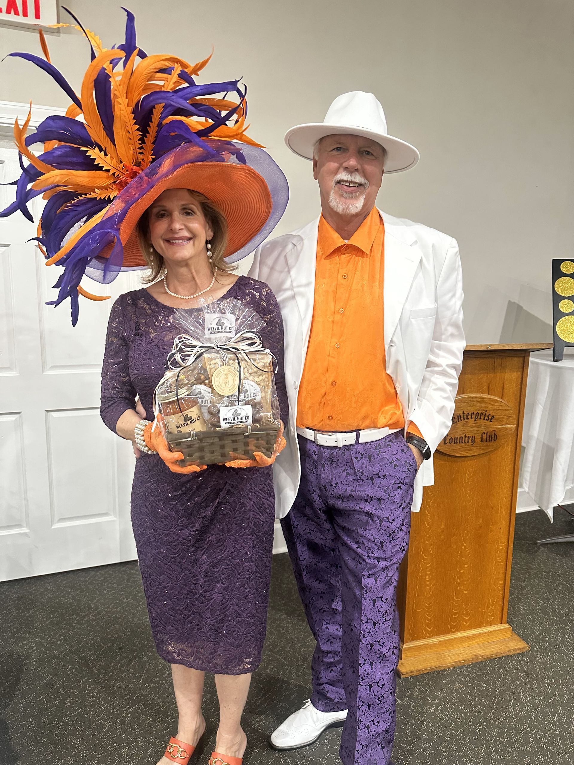 Woman in purple dress and large hat, man in white suit and purple pants, both with orange accents, pose together.