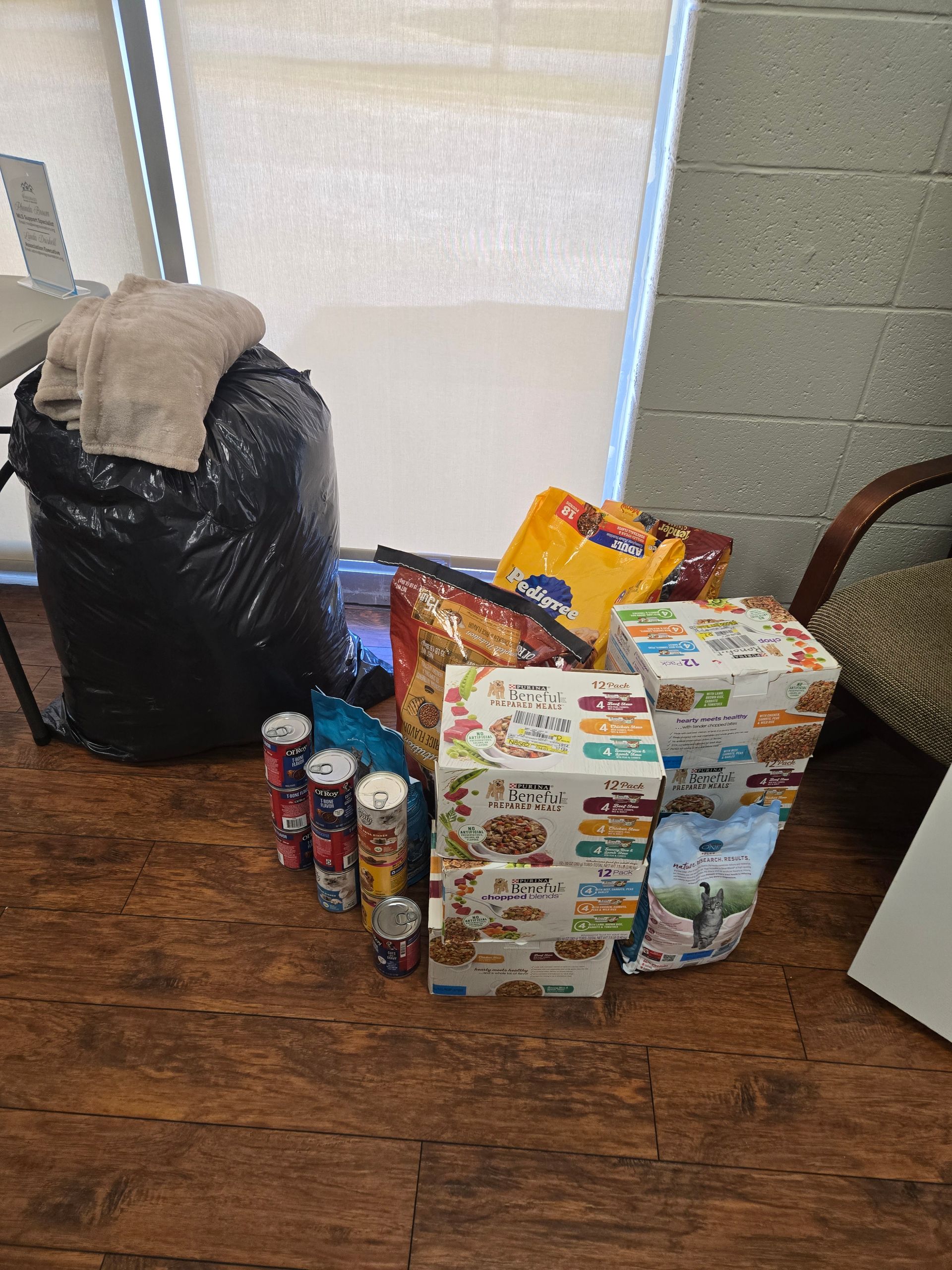 A black trash bag, boxes of food, and cans are on a wooden floor in front of a window.