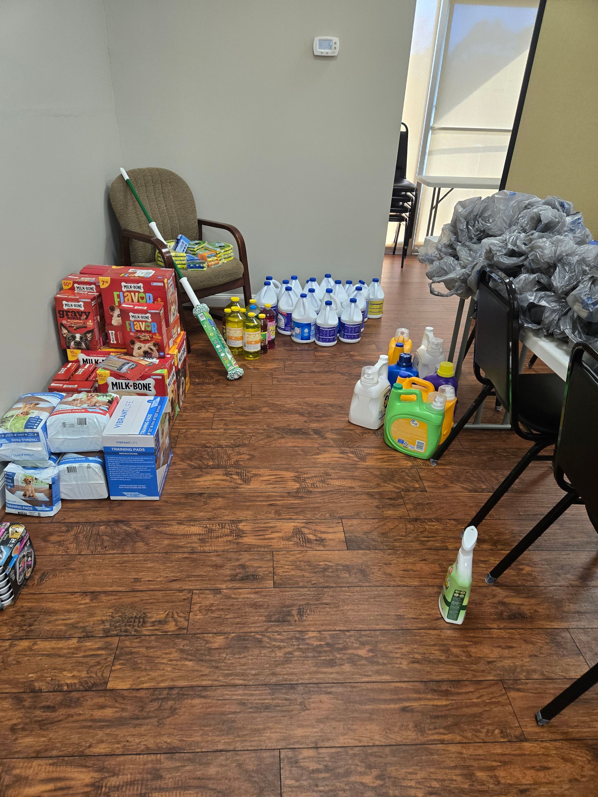 Room with boxes of diapers, cleaning supplies, toys, and a chair. Wooden floor.