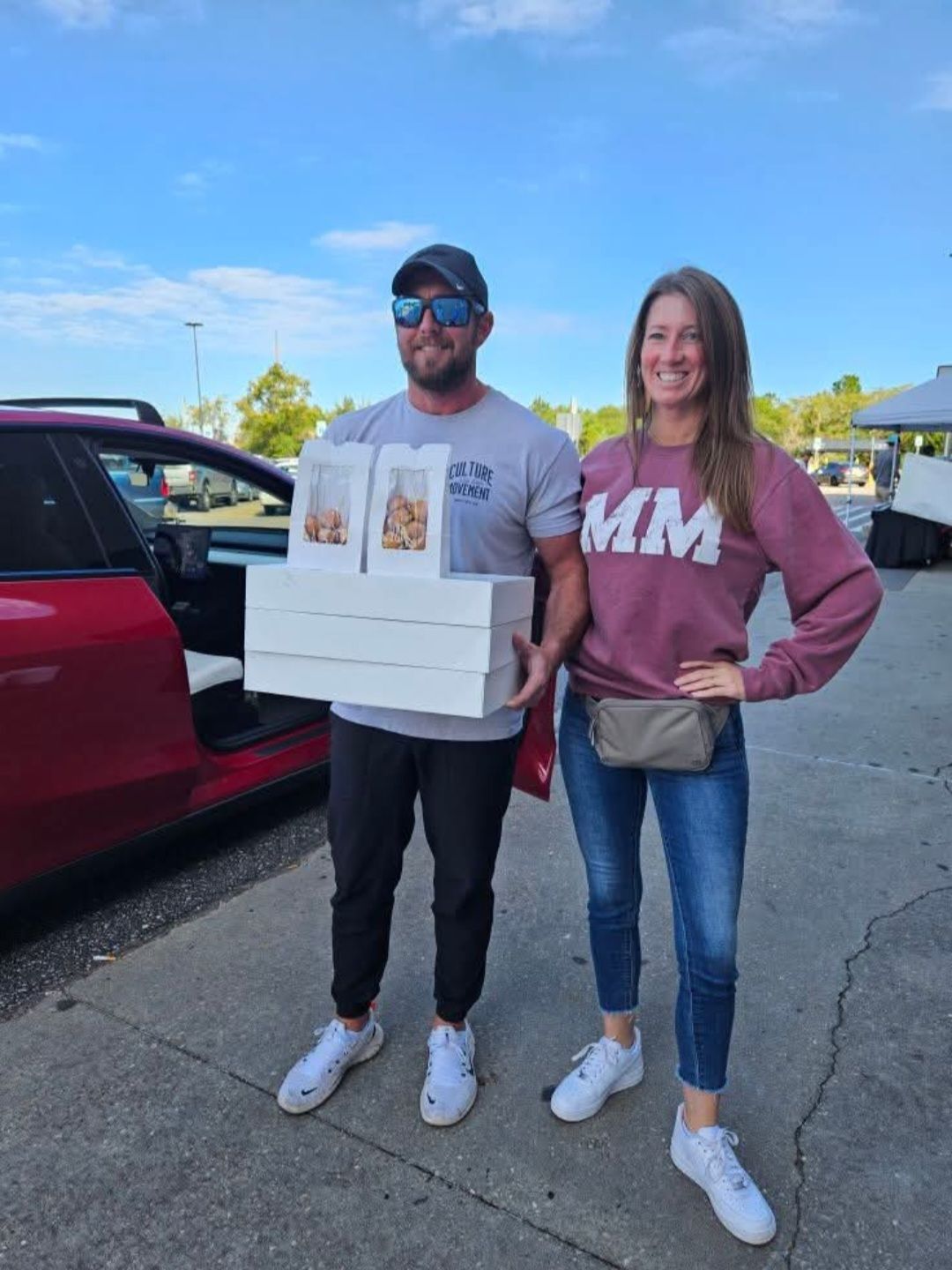 Man and woman holding boxes of baked goods near a red car, under a blue sky.