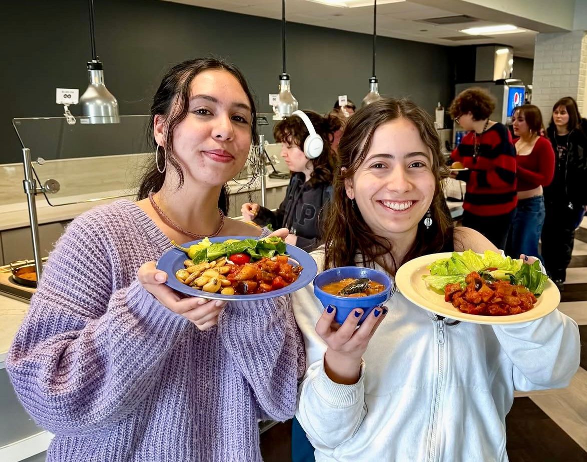Two people smiling, holding plates of food in a cafeteria. Others are in the background, near serving areas.