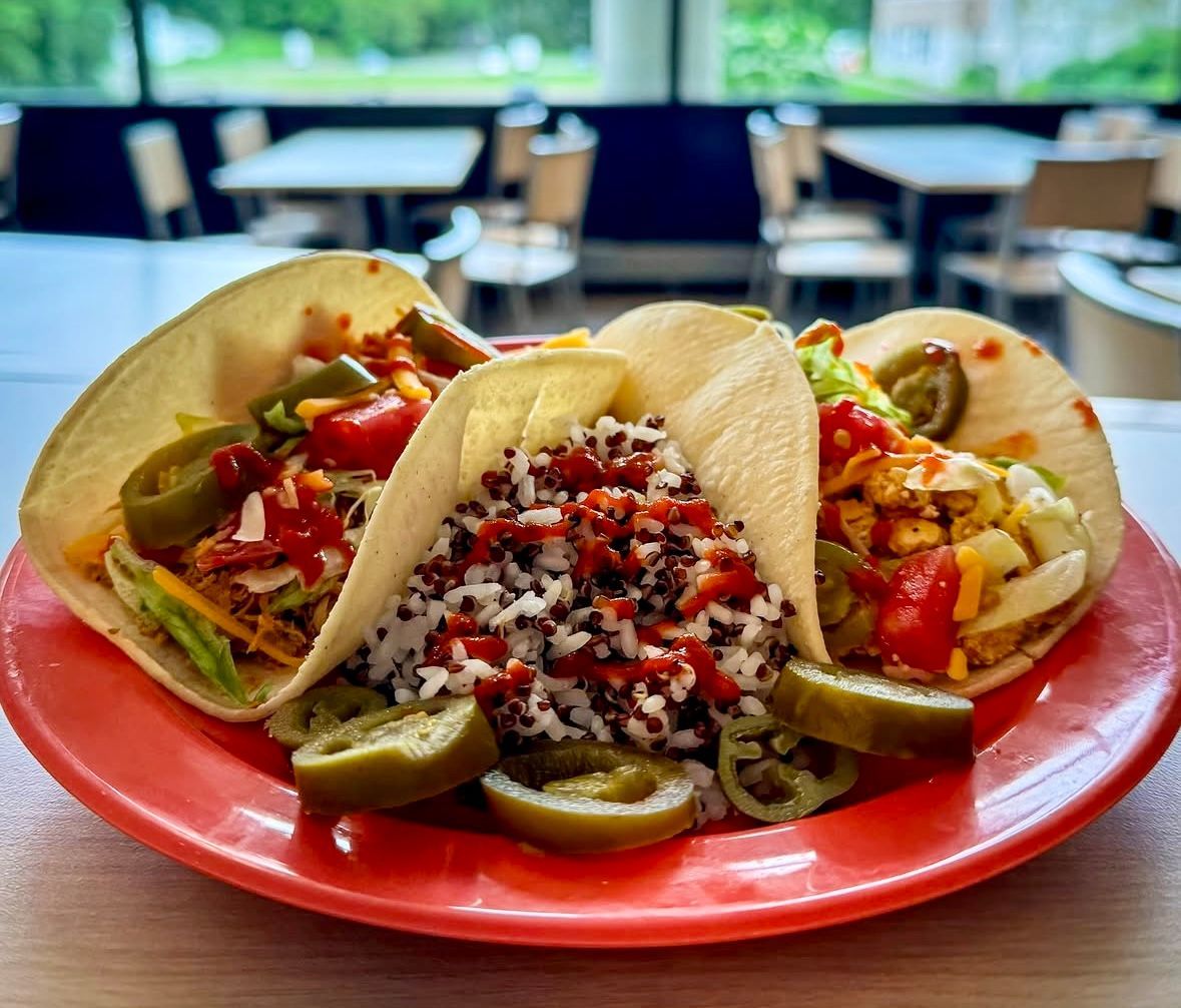 Three tacos on a red plate, filled with various ingredients, topped with pickled jalapeños, with a restaurant setting in the background.