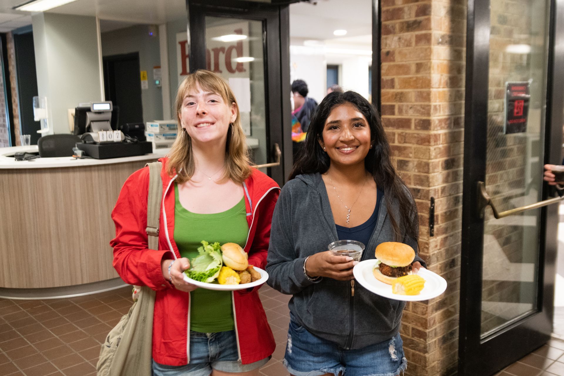 Two people smiling, holding plates of food in a cafeteria. Others are in the background, near serving areas.