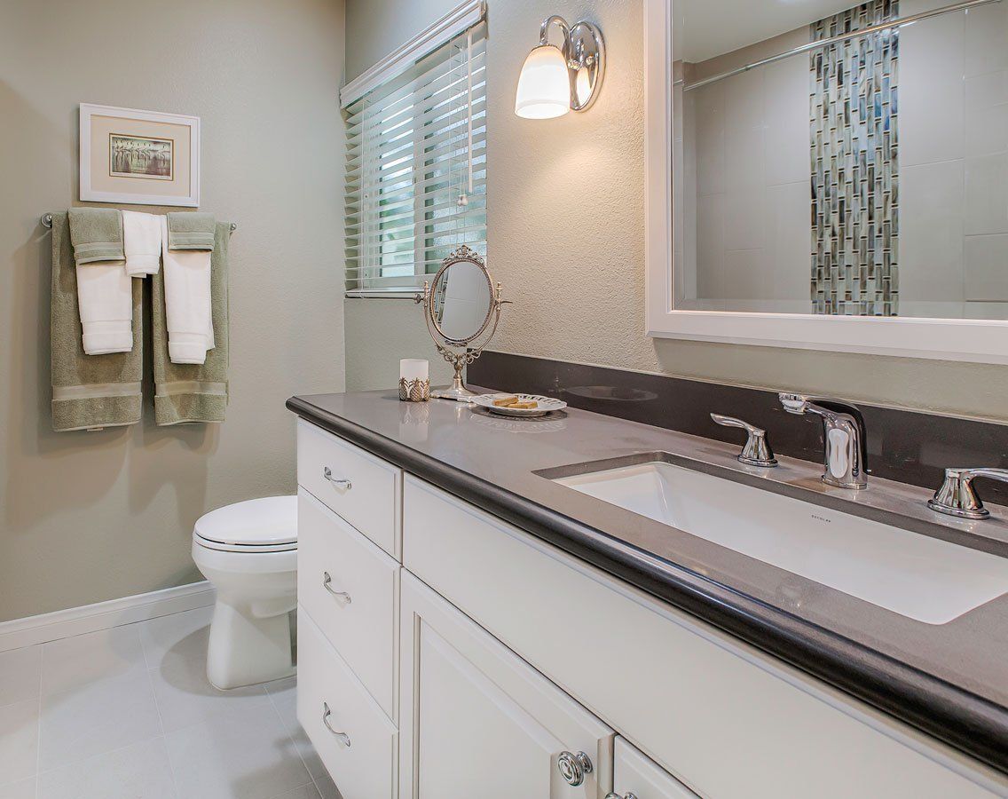 Bright bathroom with white vanity, large mirror, toilet, towel rack, and window blinds.