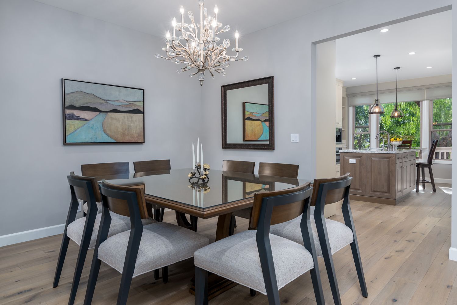 Modern dining room with a glass table, upholstered chairs, chandelier, and kitchen visible through an open doorway