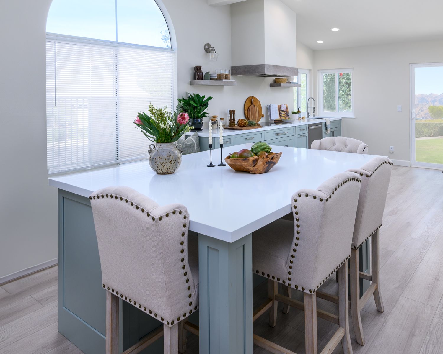Bright modern kitchen with a white island, upholstered stools, and a vase of flowers by a large window.