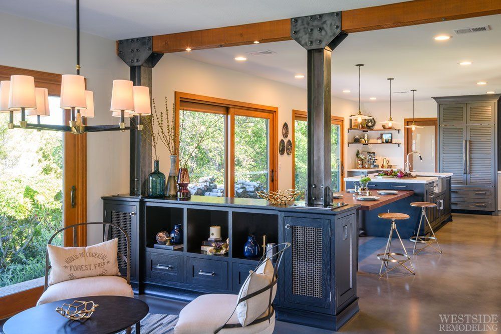 Open-plan living room and kitchen with dark wood beams, blue cabinetry, and large windows.