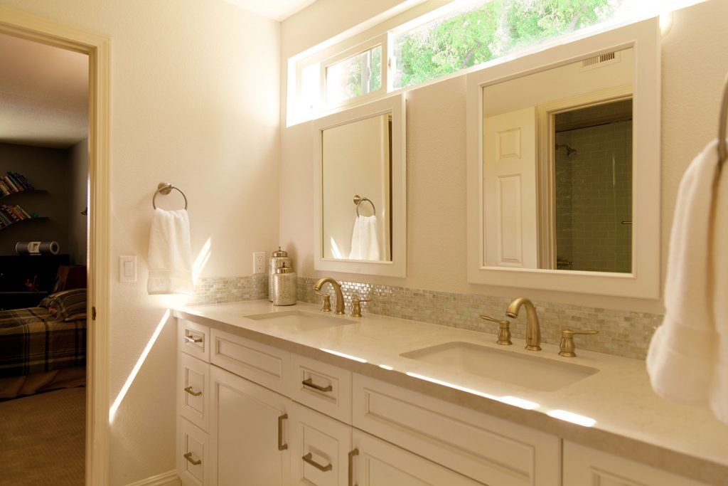 Bright bathroom vanity with double mirrors, white cabinets, and gold fixtures by a sunlit window
