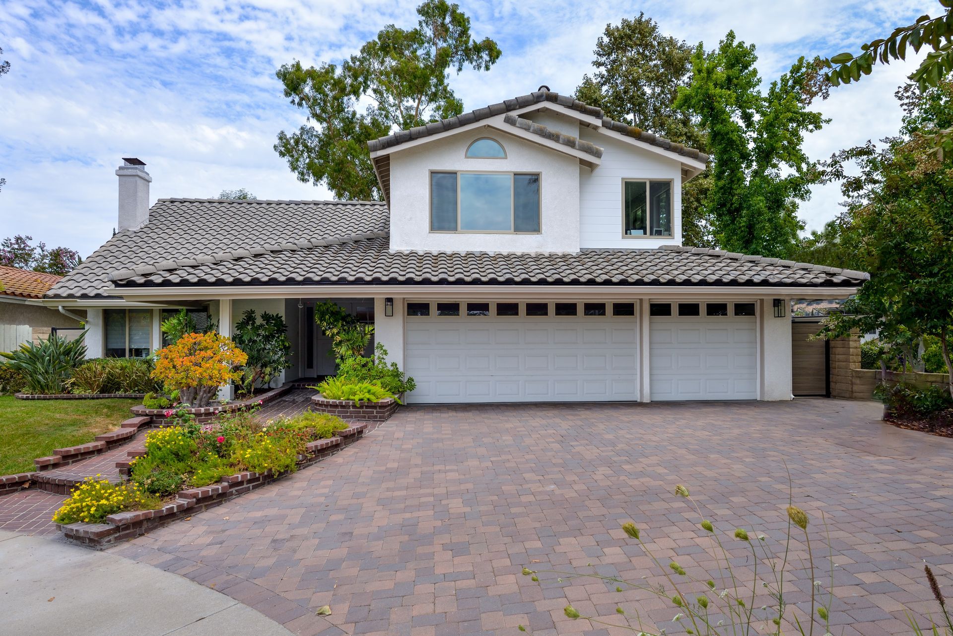 Two-story suburban house with a tiled roof, white garage doors, and a brick driveway amid lush landscaping