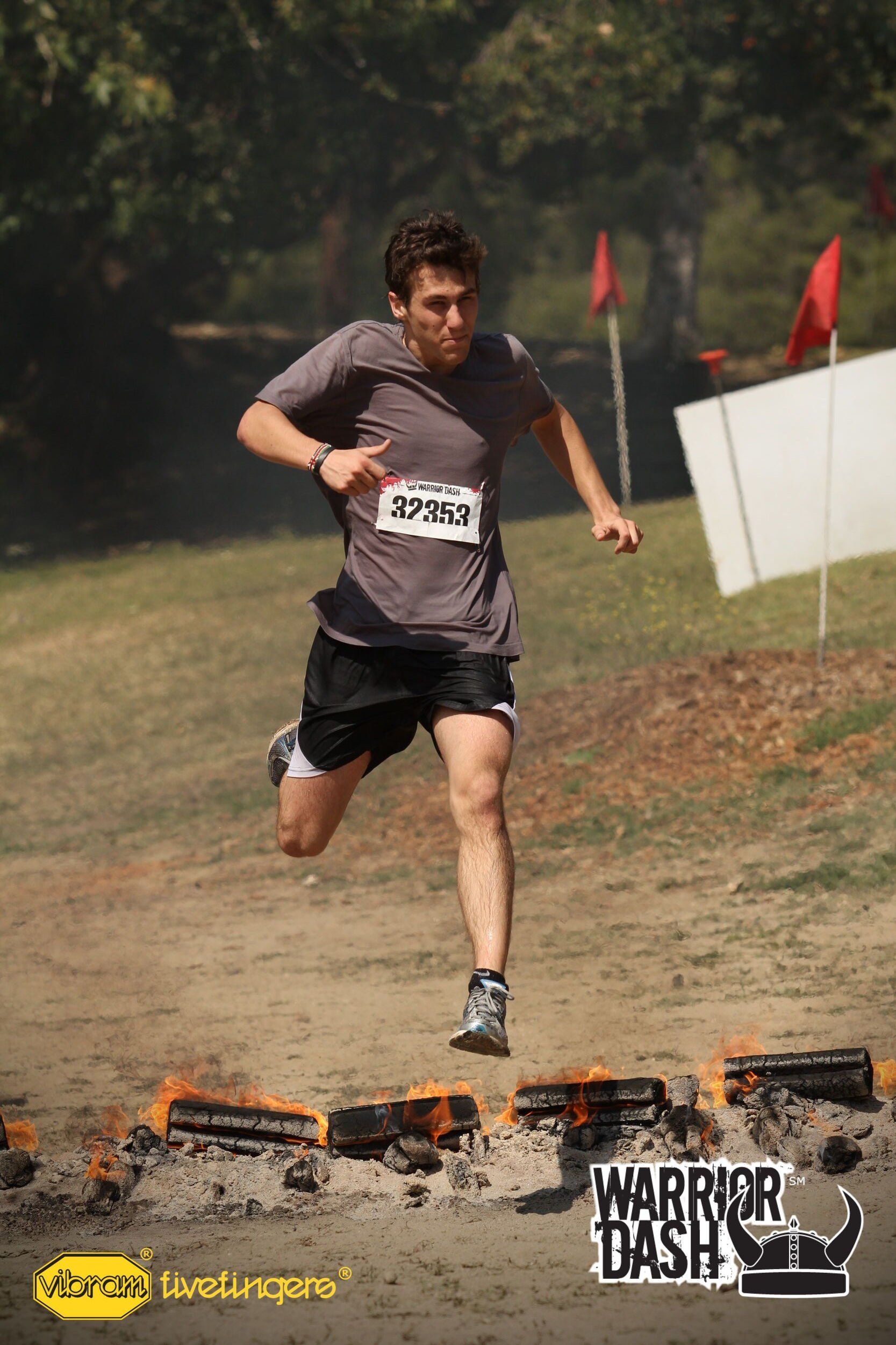 Runner jumping over flaming obstacle in an outdoor obstacle race, wearing a gray shirt and black shorts.