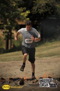 Runner in gray shirt and black shorts sprinting uphill on a dirt trail, race bib visible.