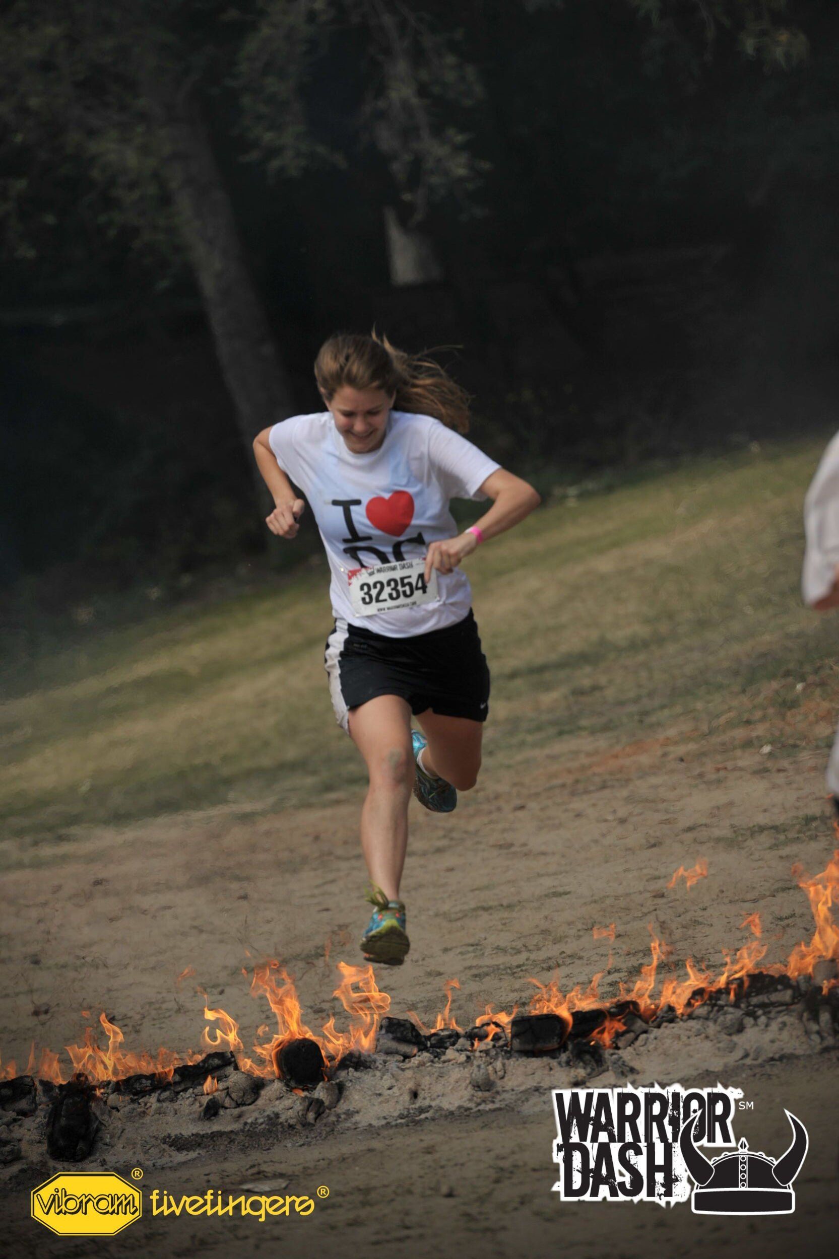 Runner in white shirt leaps over fire during an outdoor obstacle race.