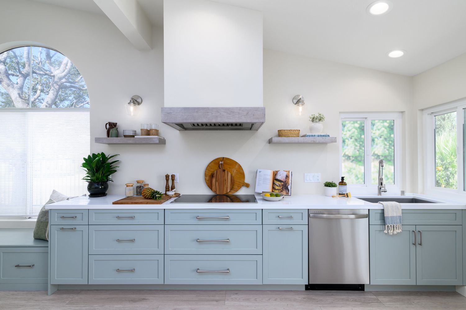 Bright white kitchen with light blue cabinets, stainless dishwasher, and arched window above the sink.