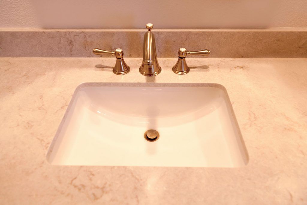 White bathroom sink with gold faucet and beige countertop under a mirror