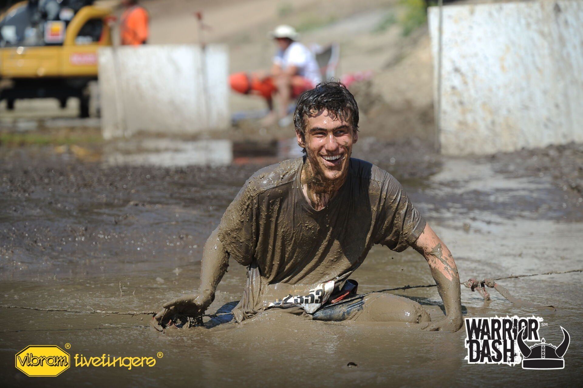 Mud-covered runner crawling through a muddy obstacle course, smiling near a barrier.