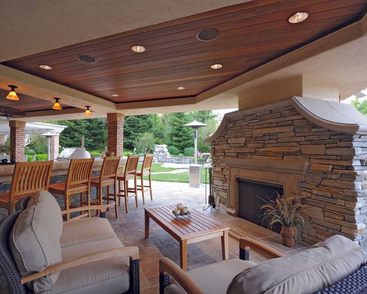 Covered patio with stone fireplace, wooden ceiling, cushioned seating, and outdoor dining area overlooking greenery