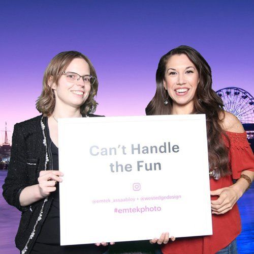 Two women holding a “Can’t Handle the Fun” sign at a purple-lit event booth
