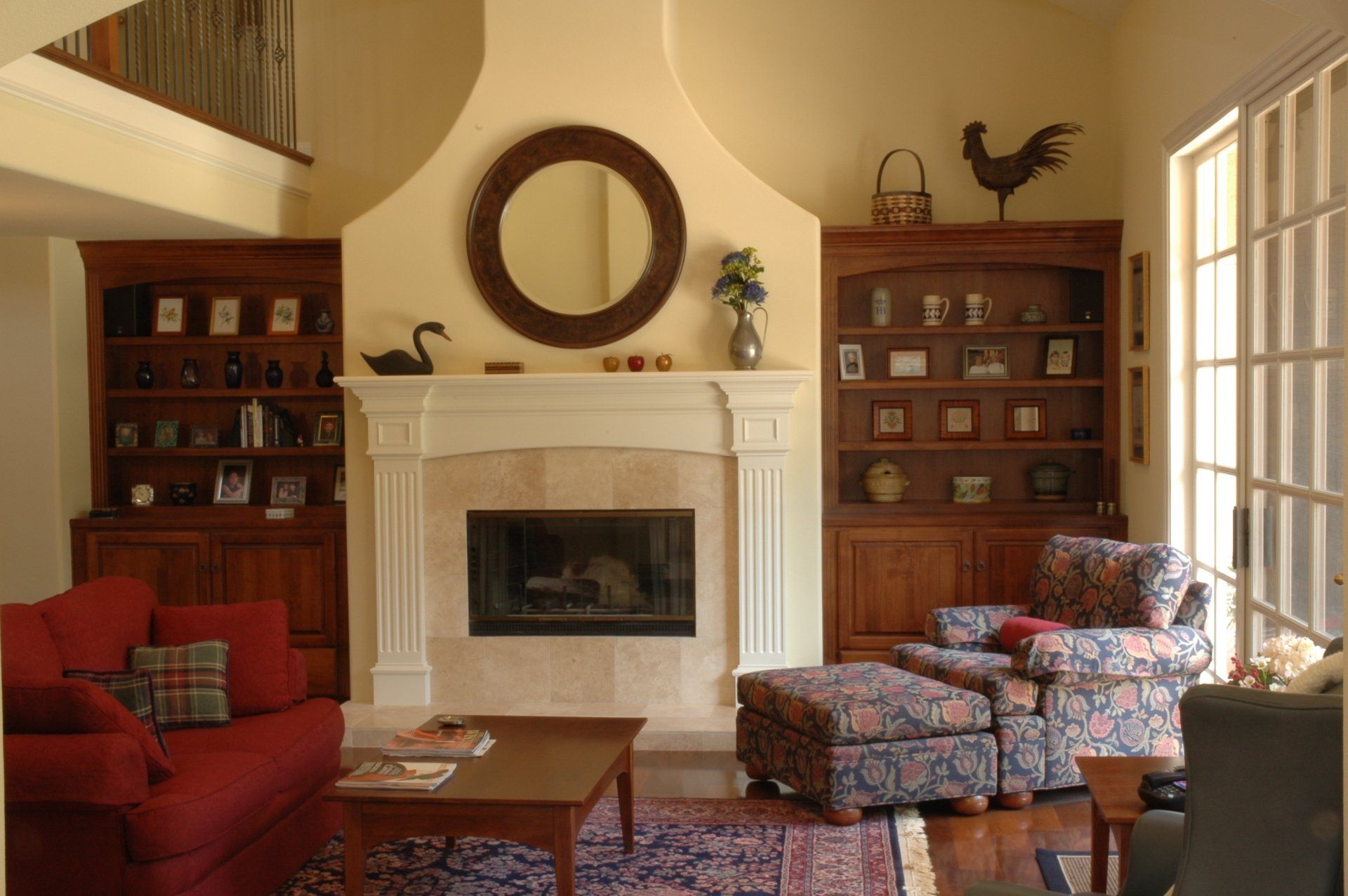 Cozy living room with a fireplace, red armchair, patterned chairs, bookshelves, and a round mirror