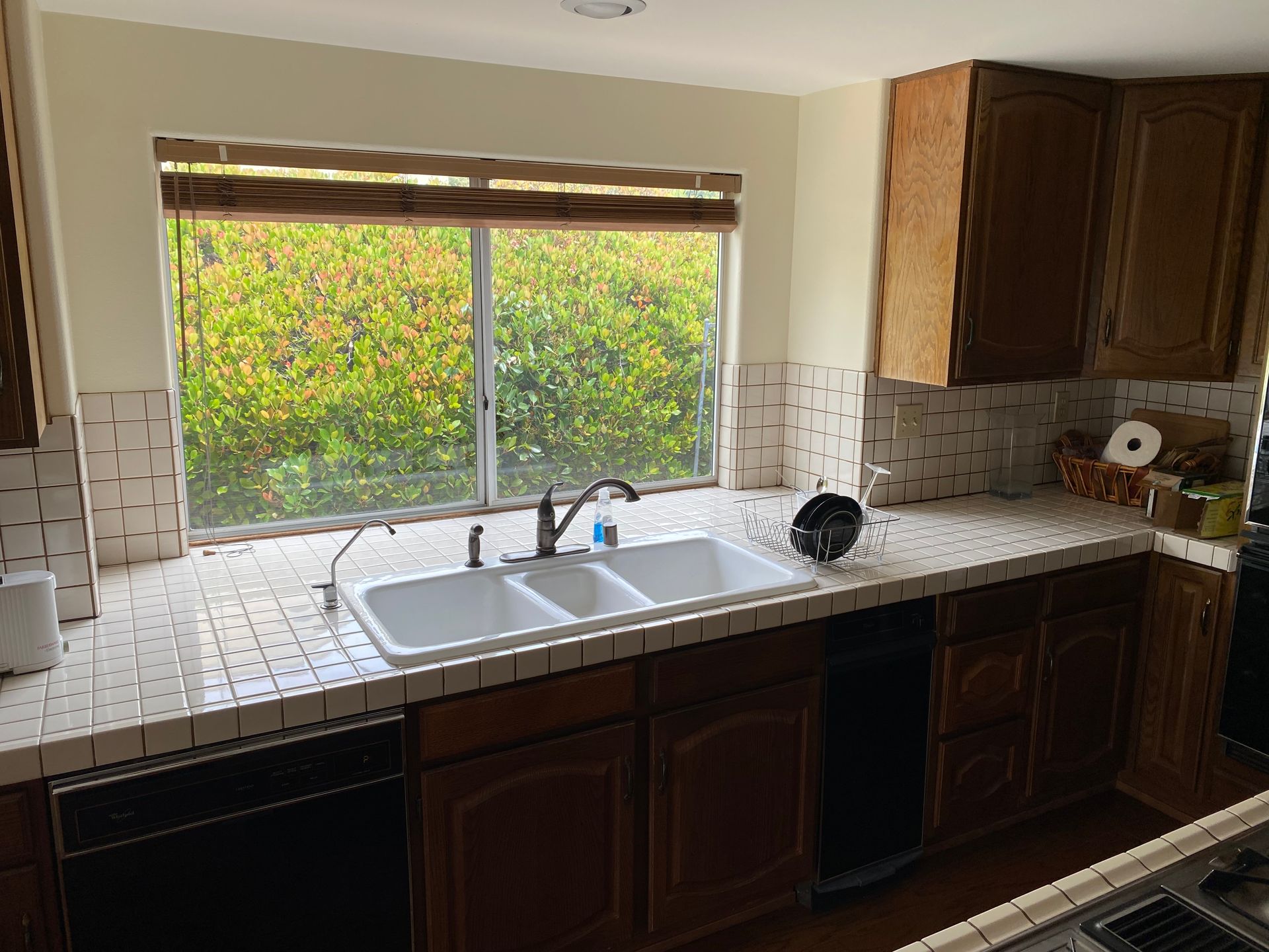 Kitchen sink beneath large window, with dark wood cabinets and white tile backsplash.