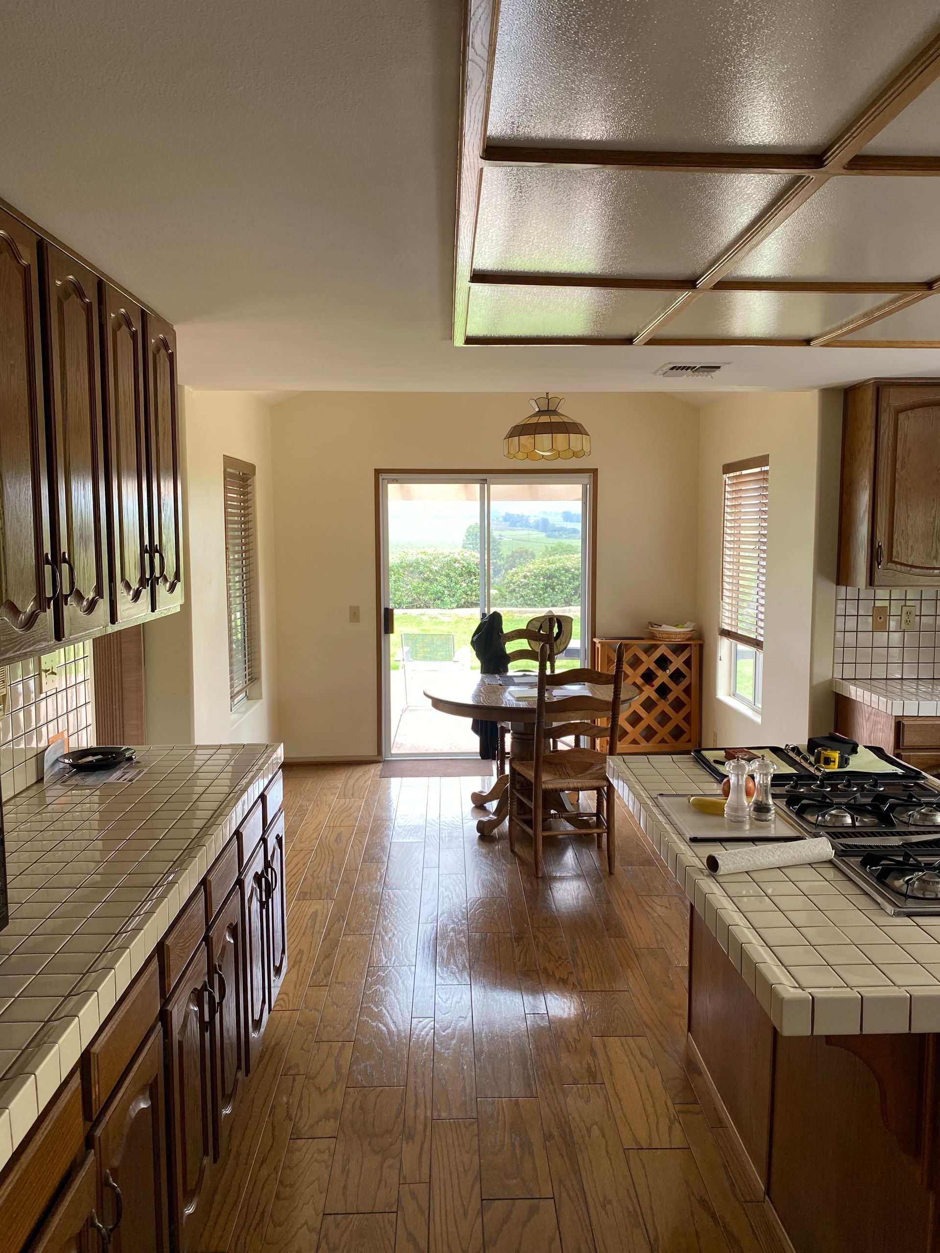 Long kitchen with wooden cabinets, tiled counters, and a bright dining nook by sliding glass doors.