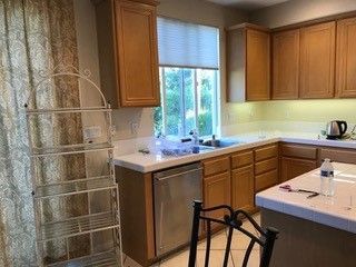 Small kitchen with wood cabinets, white countertops, sink by a window, and a metal shelving rack on the left.