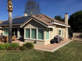 Single-story beige house with a tiled roof, green windows, and a barbecue grill on a sunny lawn.
