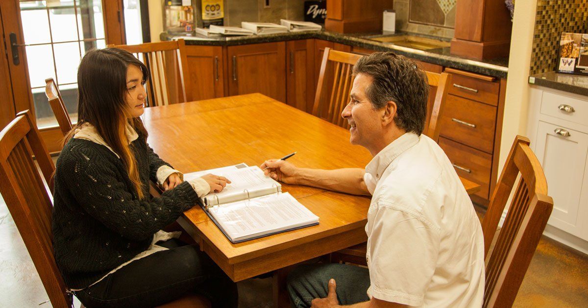 Two people sit at a kitchen table reviewing papers, with a laptop open between them.