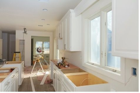 Bright white kitchen under renovation with cabinets, unfinished sink, and tools in a sunlit hallway