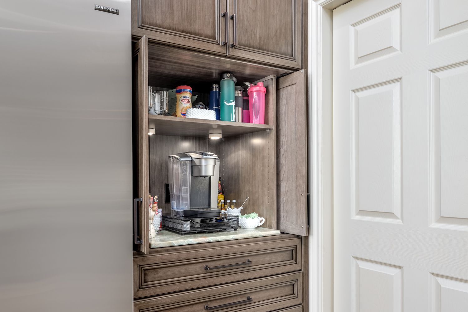 Built-in kitchen cabinet with coffee maker, mugs, and supplies beside a white door