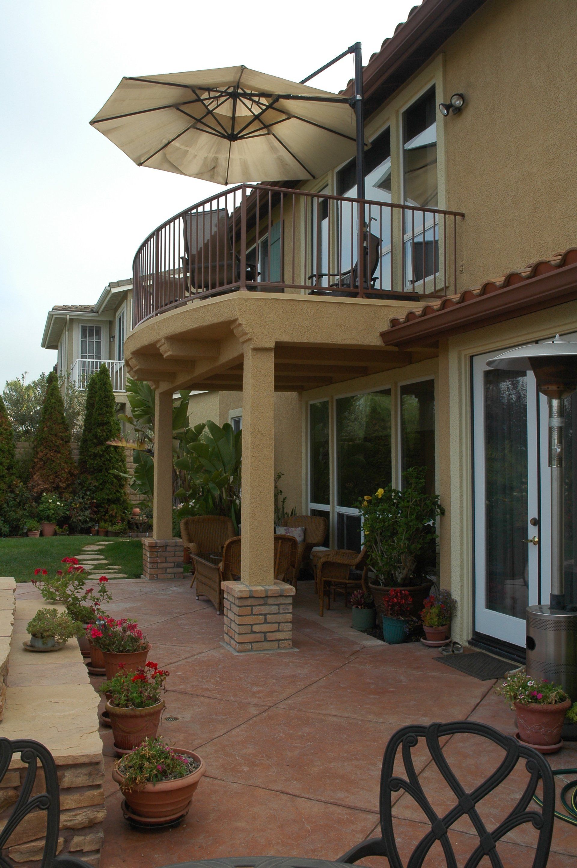 Patio with potted plants, brick floor, and a balcony with umbrella beside a stucco house.