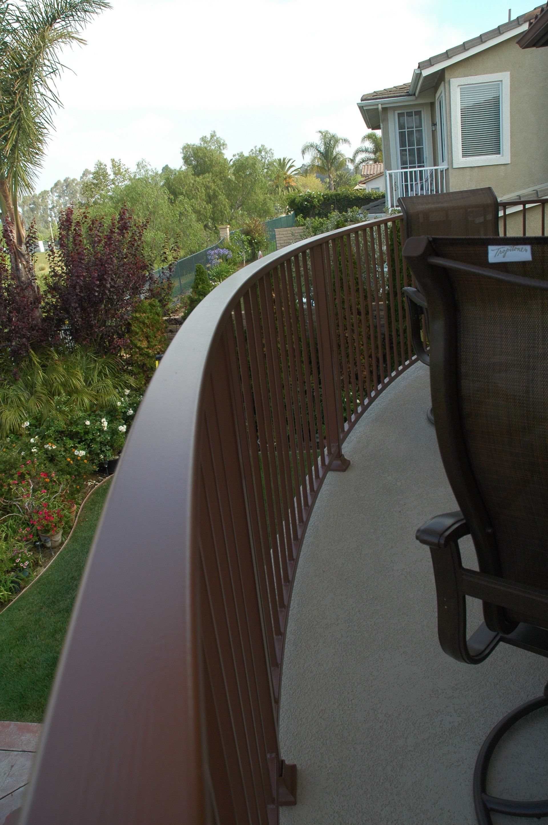 Curved balcony with brown railing overlooking a garden and house exterior