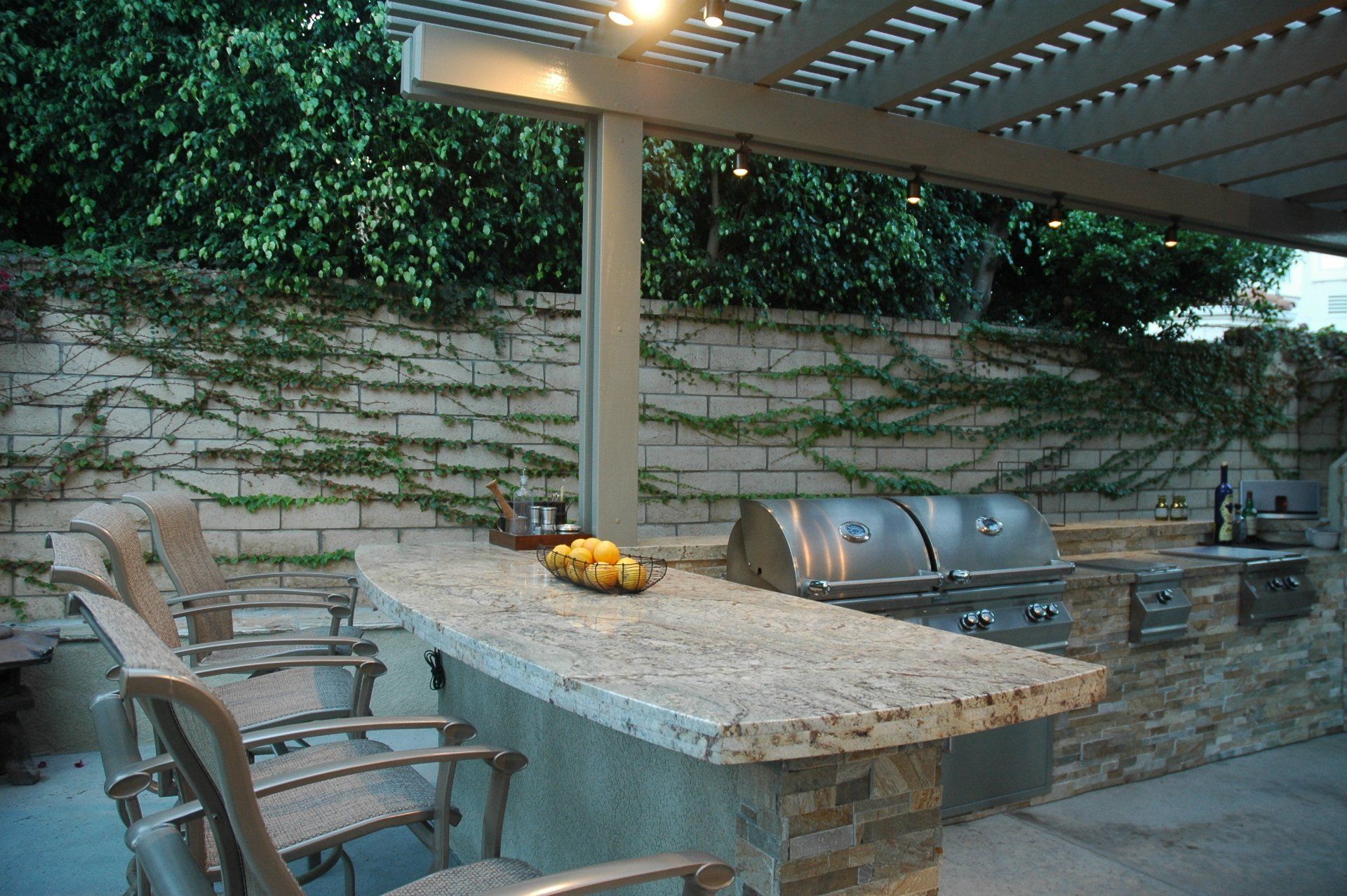 Outdoor patio with stone table, metal chairs, and built-in grill under a pergola.