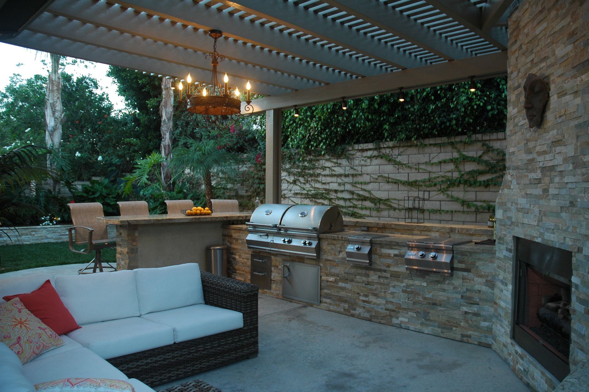 Covered patio with wicker sofa, outdoor kitchen, fireplace, and dining area at dusk