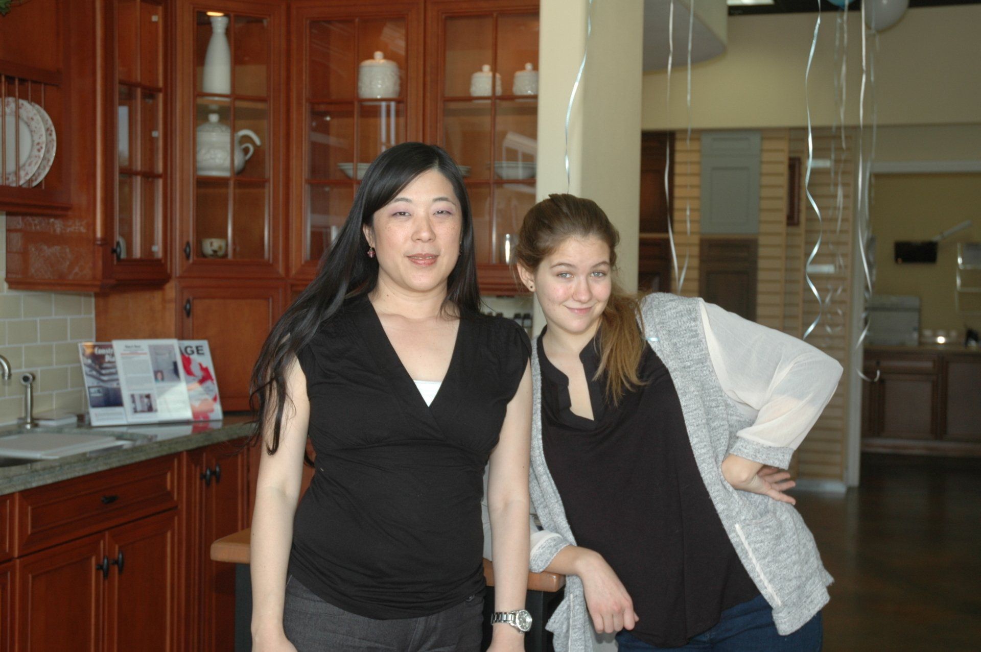 Two women posing indoors in a lobby, both wearing black tops; one holds a white bag.