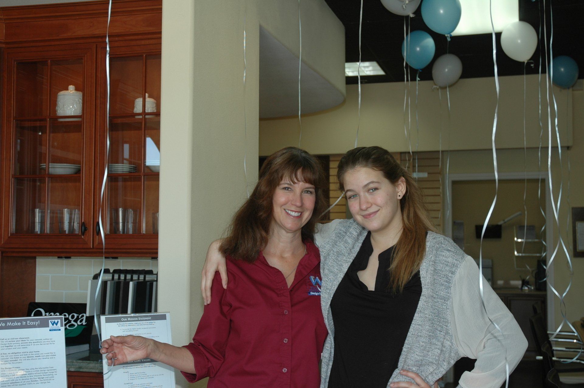 Two women smiling indoors, standing near balloons and a kitchen counter.