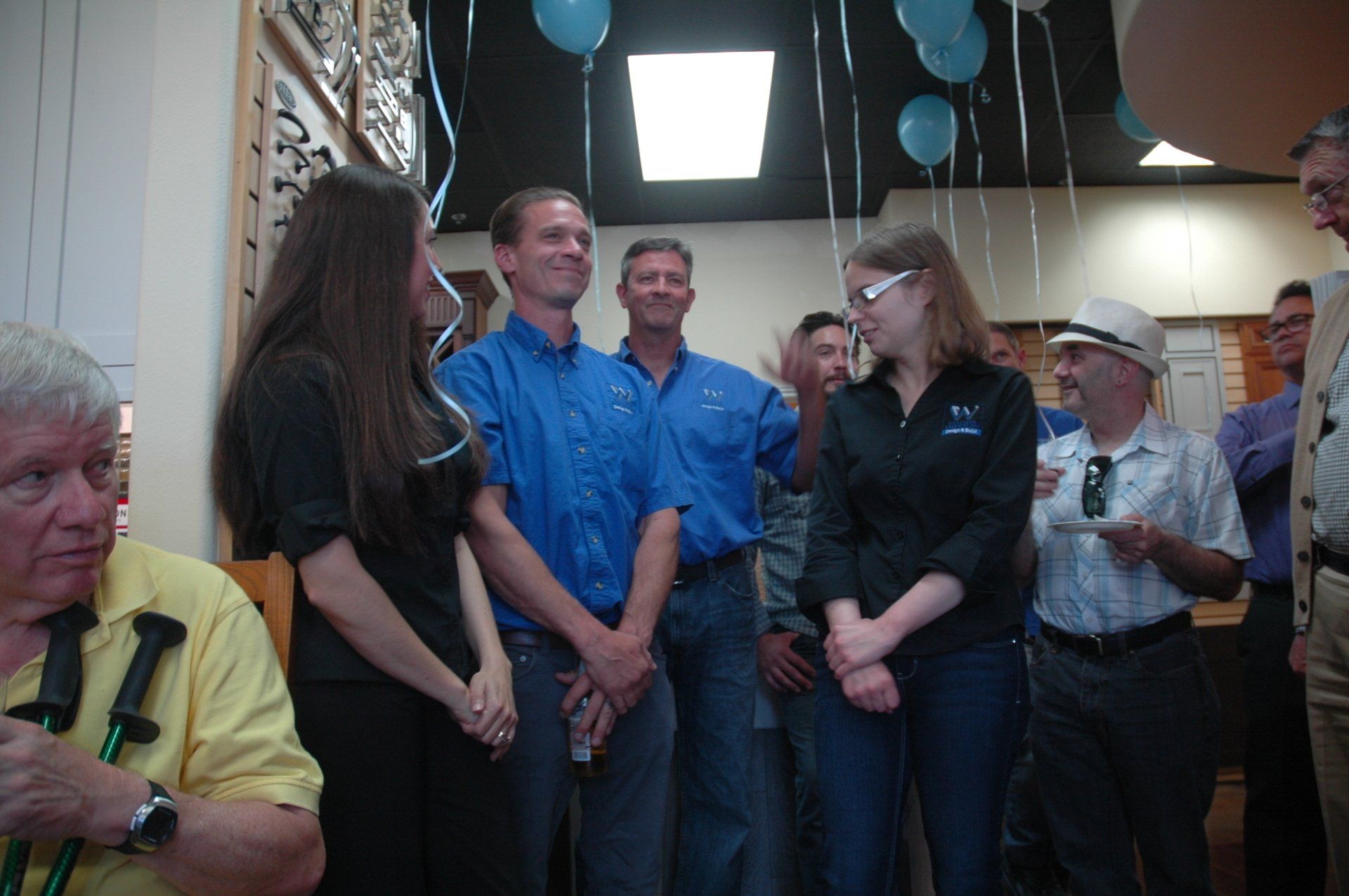 Group of people standing indoors at a gathering, talking and listening near balloons and blue hanging decorations.