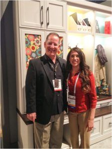 Two people standing in a kitchen showroom, smiling at the camera, with white cabinets and colorful decor.